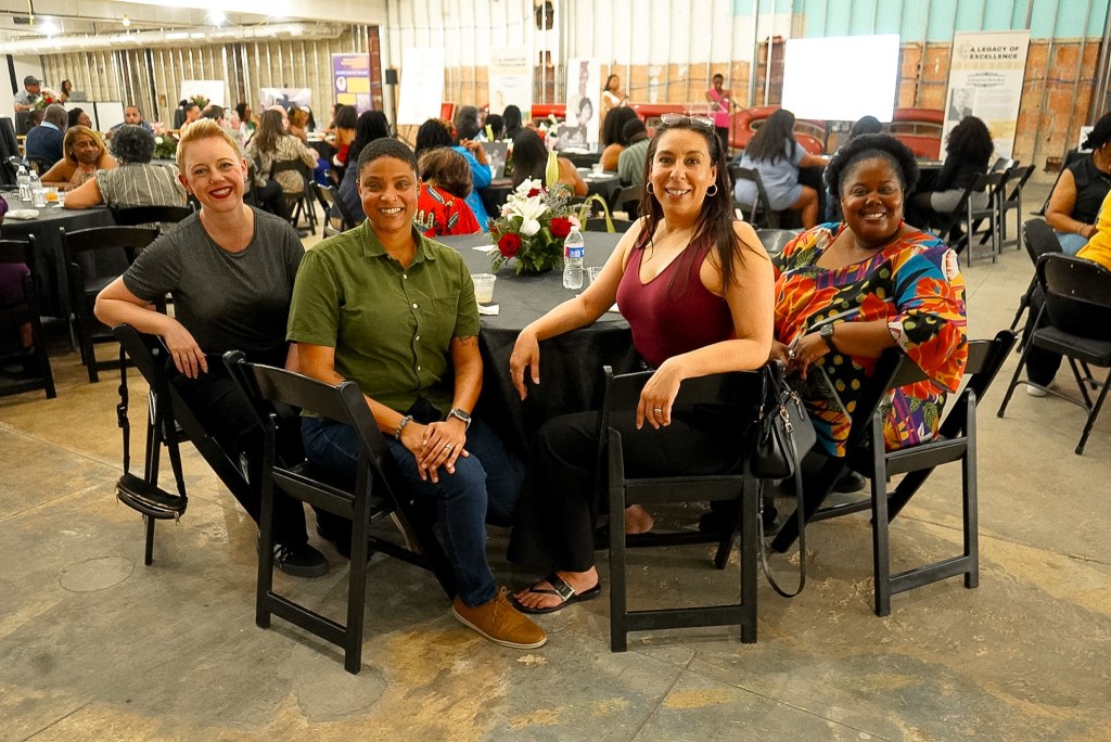 A group of four women sitting at a round table in an indoor event space, smiling and posing for the camera. The background shows other attendees mingling and engaging in discussions.